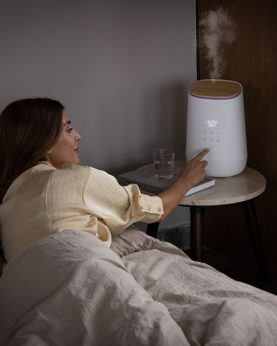 Eine Frau liegt im Bett und bedient einen eingeschalteten Luftbefeuchter auf dem Nachttisch, der Wasserdampf abgibt.